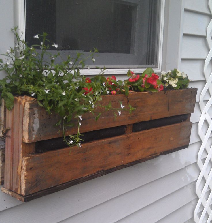 Jardinière en palettes en bois usé marron avec différentes fleurs blanches et rouges dedans. Mur blanc et fenêtre au-dessus.