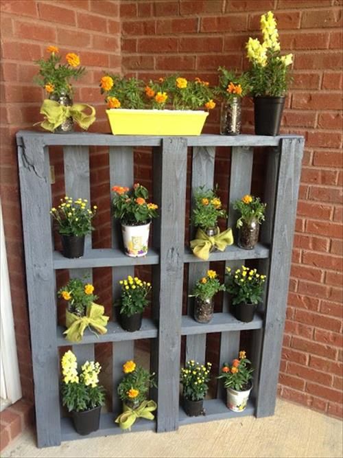 Jardinière palettes en bous gris-bleu clair avec une multitude de pots dessus et un mur en briques rouges derrière. Petites fleurs oranges et jaunes.