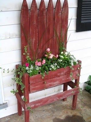 Jardinière en palettes d'une couleur rouge usé avec différentes plantes, géraniums et fleurs dedans. Mur extérieur blanc avec fenêtre noire sur le côté droit.