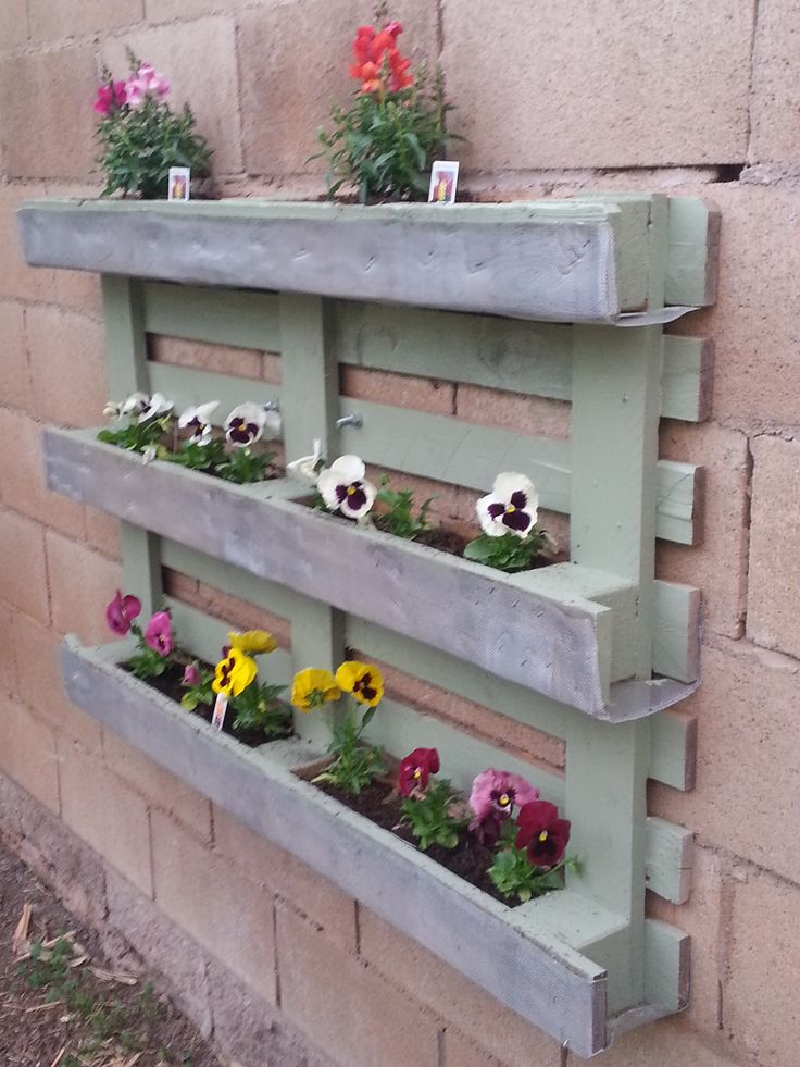 Jardinière en palette de couleur bleu-clair sur un mur beige avec dalles. Fleurs de différentes sortes dessus et de diverses couleurs comme le blanc, le jaune ainsi que le violet. jardinière en palette 1