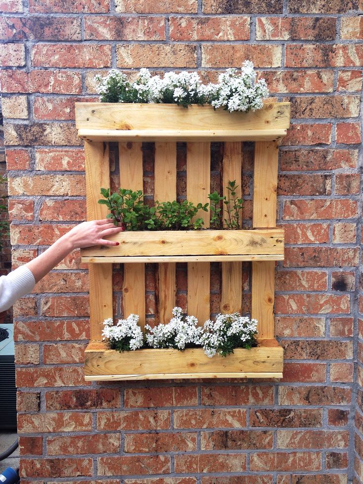 Jardinière en palette en bois hêtre à trois niveaux avec fleurs blanches dedans et plantes au milieu. Mur ancien en briques rouges.