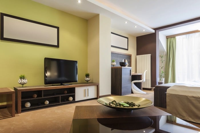 Chambre moderne aux couleurs vert clair et bois marron. Meuble TV rectangulaire en bois, grand miroir ainsi qu'une table en verre et en teck au premier-plan.