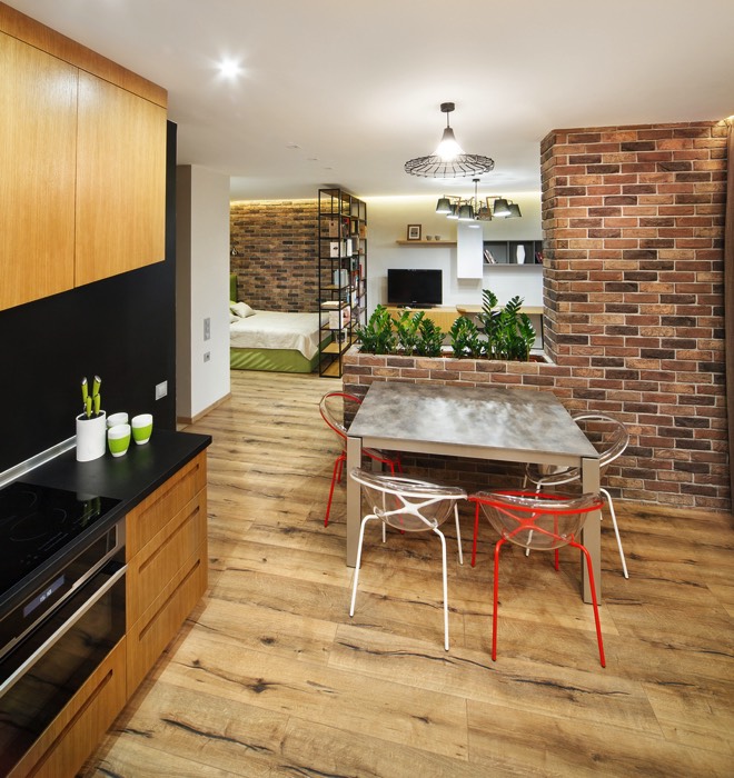 Murs en briques rouges coupé pour disposer un parterre de plantes. Plan de travail anthracite et en bois sapin sur parquet avec différentes pièces ouvertes comme une chambre et un salon.