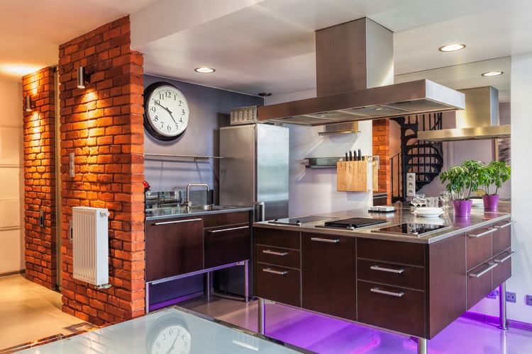 Mur en briques rouges avec spots lumineux derrière une cuisine pop moderne avec parterre violet et multitude de rangements. Grande hotte en acier, îlot central carré et grande horloge blanche.