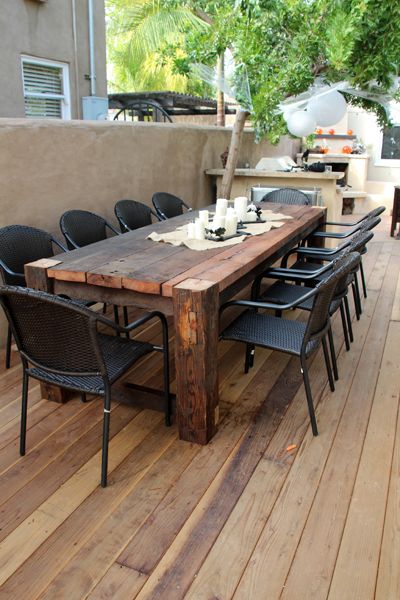 Table rectangulaire en bois teck avec six chaises noires sur parquet classique. Salle à manger extérieure au style campagnard/rustique avec mur beige servant de délimitation.