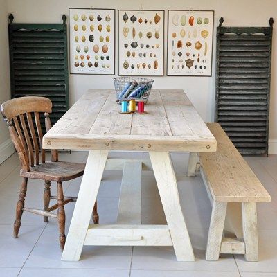 Table en bois frêne avec banc dans une salle à manger rustique. Carrelage blanc, trois tableaux blancs avec fruits et légumes dessus, deux portes style ancien des deux côtés.