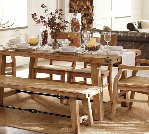 Table rectangulaire en bois hêtre au style campagnard/rustique avec banc sur tapis beige uni. Pièce avec mur blanc, salon marron sur la droite et stock de bois au fond.