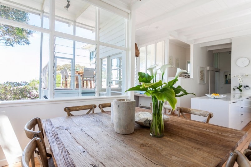 Salle à manger au style campagnard/rustique avec table carré en bois et fleurs blanches dessus. Cuisine blanche sur la droite, couloir au fond et grande baie vitrée à gauche.