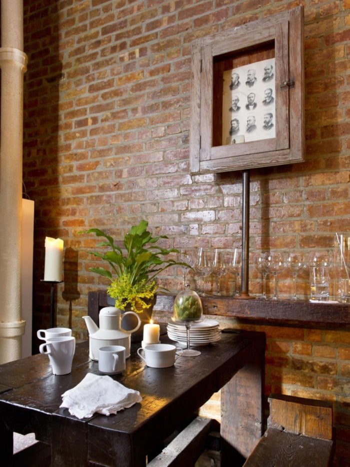 Mur en briques au-dessus d'une petite table à manger en bois wengé avec bancs. Rangement avec différents verres dessus, bougie, tableau avec photos de personnes noir et blanc dedans.