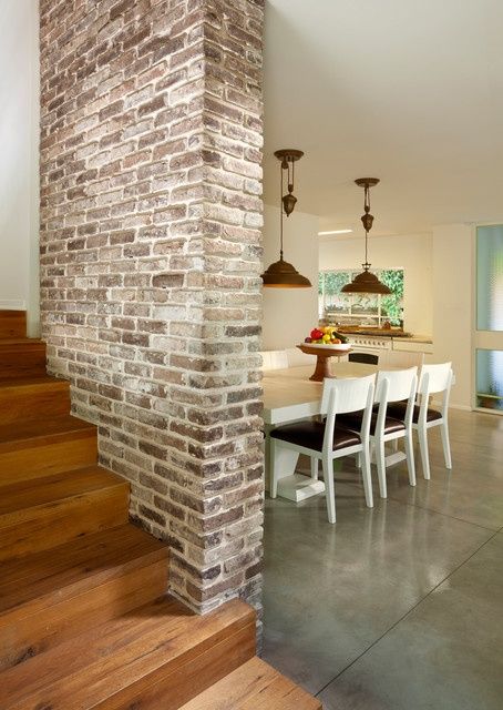 Mur en briques beiges, blanches et marrons servant de séparation entre un escalier en bois ainsi qu'une salle à manger. Table blanche, chaises avec coussins violet foncé, avec deux lampes suspendues au-dessus.