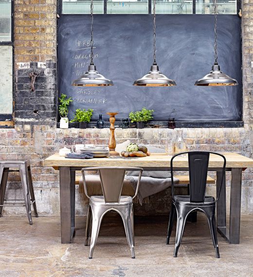 Salle à manger avec trois lampes en inox suspendues au-dessus d'une table en bois. Mur en vieilles briques et tableau noir rectangulaire avec inscriptions à la craie dessus.