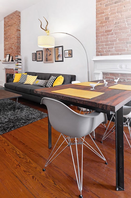 Pièce blanche avec mur blanc doté de deux cheminées en briques rouges ainsi que d'un parquet stratifié. Pièce au style industriel avec table, canapé noir, coussins jaunes et tapis.