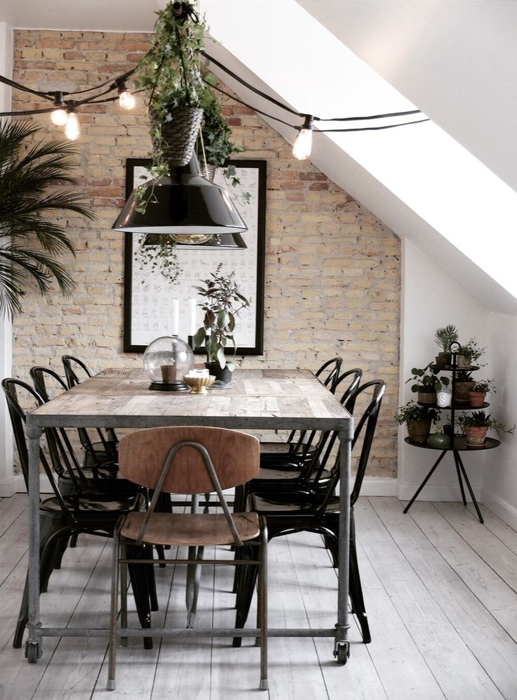 Salle à manger style industriel avec table, chaises noires sur parquet. Mur en briques beiges avec deux lampes noires suspendues dans une pièce légèrement green power.