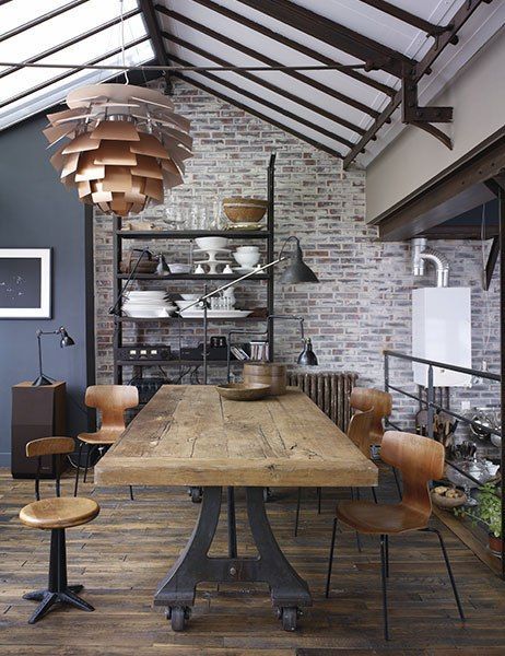 Salle à manger avec style industriel munie d'une table en bois avec chaises. Luminaire en bronze original, mur en vieilles briques rouges et étagère noire avec notamment assiettes dessus.