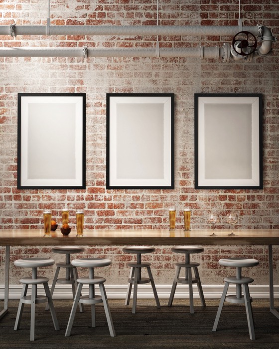 Salle à manger style industriel avec mur en briques rouges et trois tableaux rectangulaire gris, blancs et noirs. Table en bois avec tabourets de bar rondes.