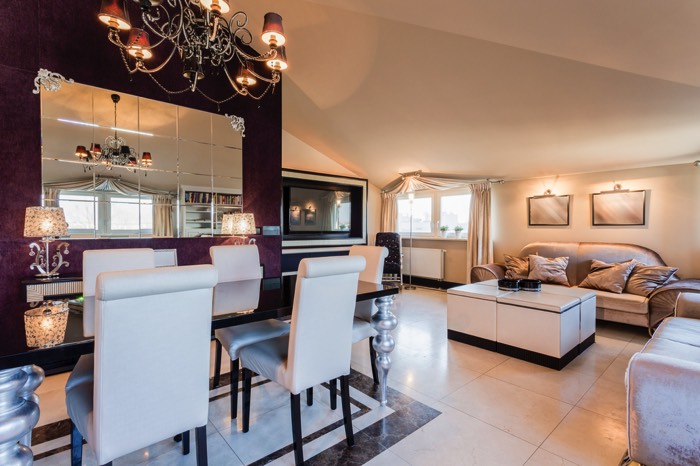 Décoration pour moderniser une salle à manger traditionnelle blanche avec longue table noire, chaises blanches, salon avec grande TV. Grand miroir sur mur violet et lustre.