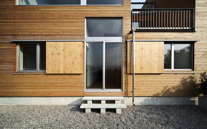 Aménagement de graviers autour d'une maison en bois style chalet. Fenêtres originales avec fermetures coulissantes et balcon à l'étage.