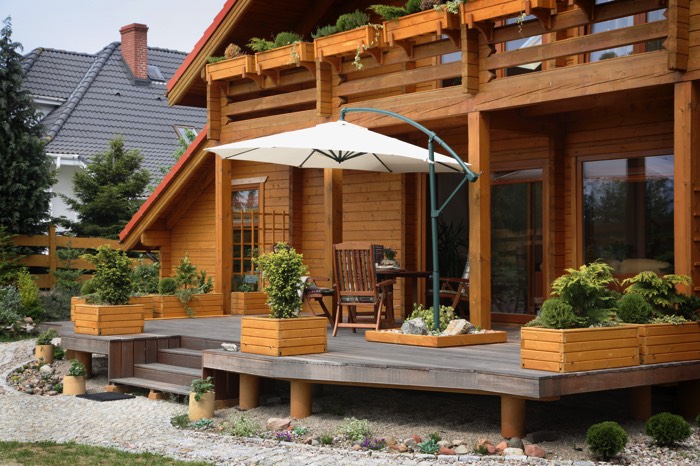 Disposer du parterre de gravillons tout autour de la maison. Habitation style chalet en bois cerisier avec terrasse doté d'un parasol blanc et d'un petit salon extérieur.