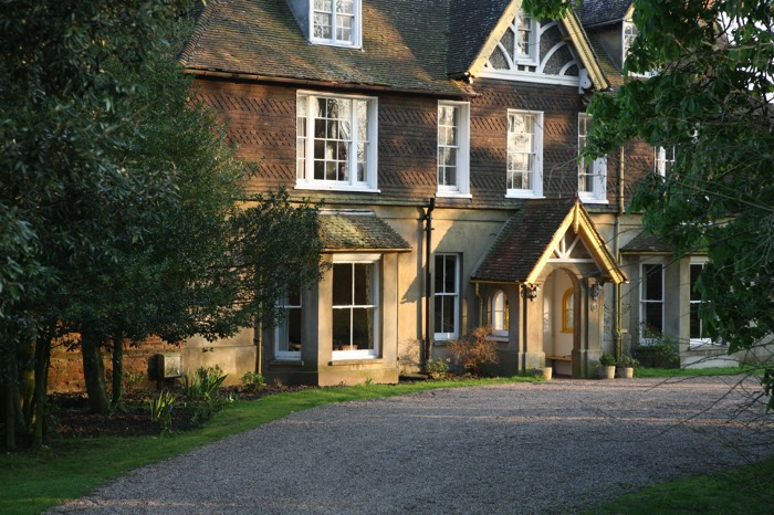 Aménagement d'une entrée gravillonnée bordant une pelouse avec différents arbres. Maison à étages style rustique avec fenêtres blanches.