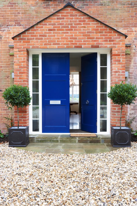 Aménager une entrée gravillonnée devant une maison en briques rouges. Double porte de couleur bleue claire avec deux petits arbustes à l'entrée.