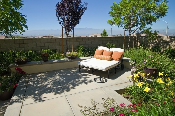 Aménagement d'une terrasse zen avec grandes dalles en béton. Environnement green power avec fleurs jaunes, rouges et lit blanc et orange.