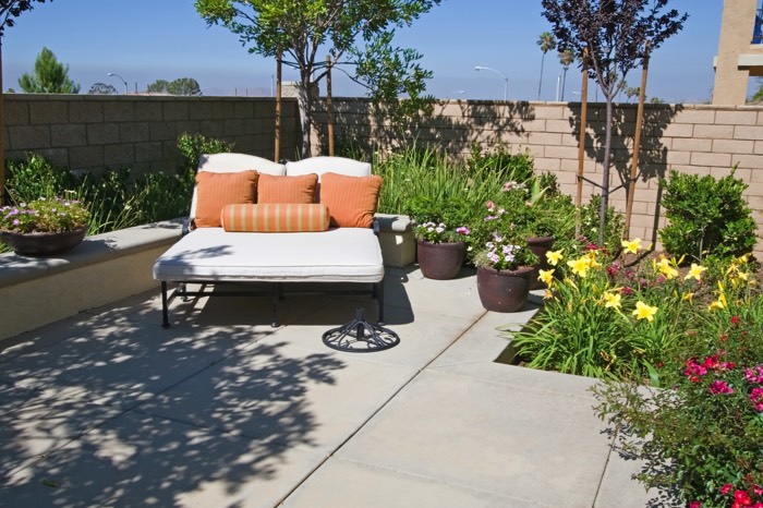 Terrasse zen avec lit deux places blanc et coussins oranges. Multitude de plantes et d'arbustes autour avec mur non peint en parpaings.