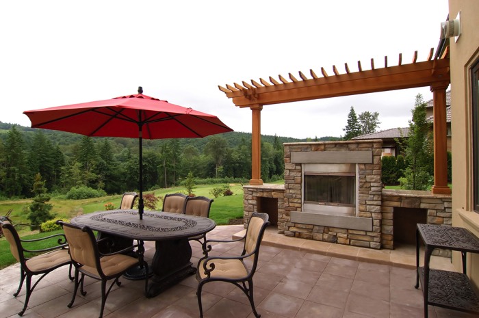 Aménager une terrasse zen avec une pergola en bois cerisier abritant un barbecue en pierres. Carrelage marron avec magnifique table ovale, parasol rouge et sièges beiges.