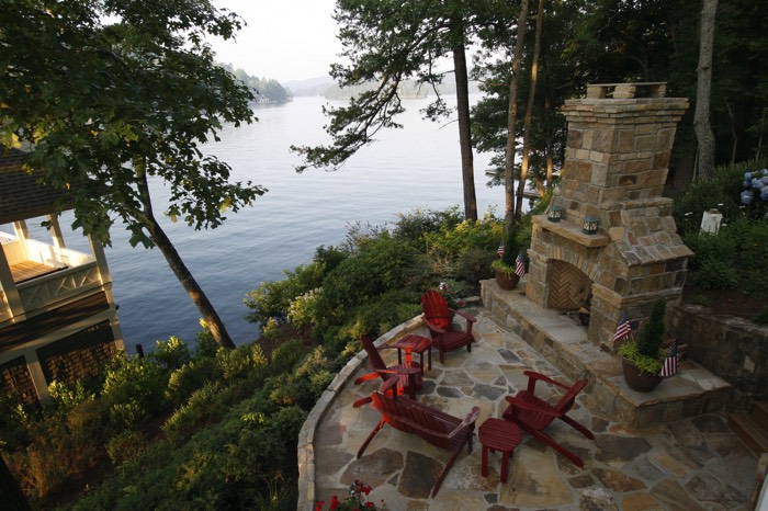 Aménagement d'une terrasse zen avec panorama sur un lac entouré d'une forêt. Vieille cheminée en pierres avec petit salon extérieur en rouge.