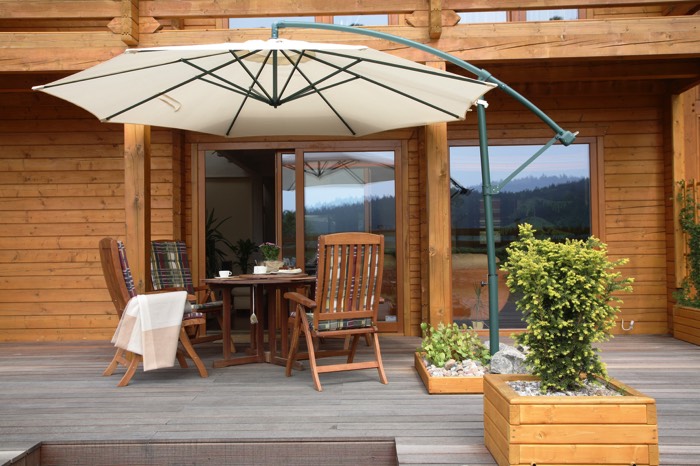 Aménager une terrasse zen en bois hêtre et bouleau avec parquet. Grand parasol blanc abritant un salon extérieur en bois avec baie vitrée derrière.
