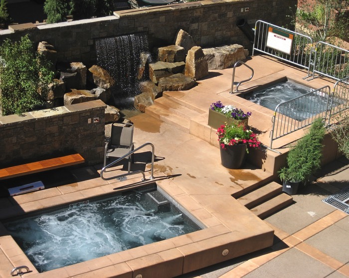 Aménagement d'une terrasse zen en dalles beiges avec spa et petite piscine rectangulaire. Magnifique fontaine en pierres, banc en bois et plantes.