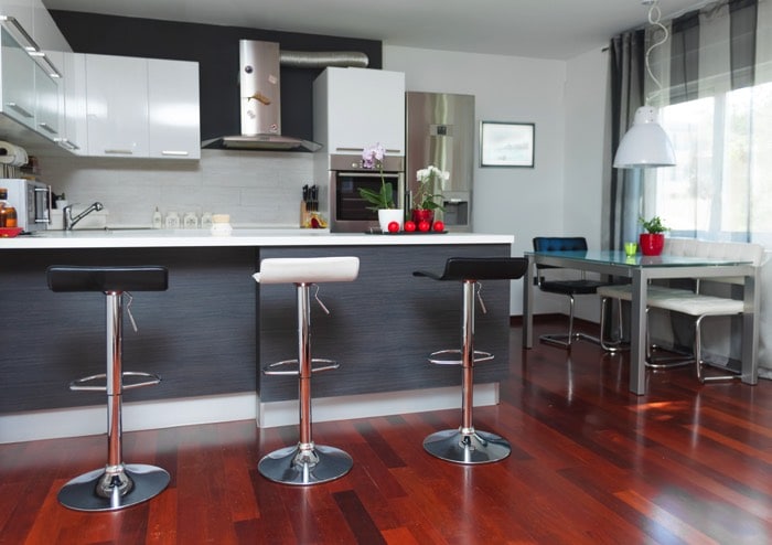 Cuisine gris avec bar blanc sur magnifique parquet stratifié. Table à manger en verre, sièges de bar, frigo blanc et hotte en acier.