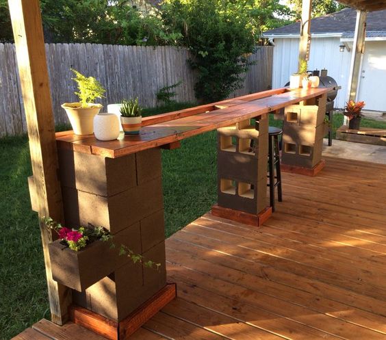 Parpaings décoratifs servant à installer un bar en bois noyer dans le jardin, avec tabourets noirs. Pergola, parquet et clôture.
