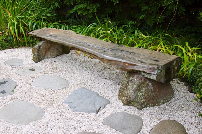 Banc rustique en bois avec deux gros cailloux servant de support pour décorer son jardin. Végétation en arrière, sable et gros cailloux servant d'allée.