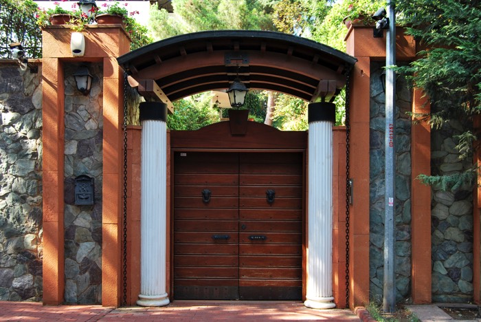 Entrée de portail avec magnifique arche en bois wengé style zen. Deux colonnes blanches, colonnes oranges avec grand mur en pierres.