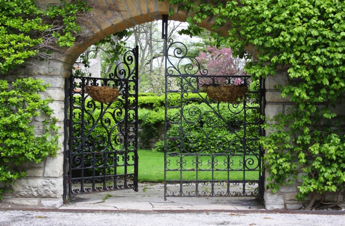 Entrée portail avec grand arche en pierres style ancien et plantes grimpantes. Portail noir à double battants avec de belles gravures.