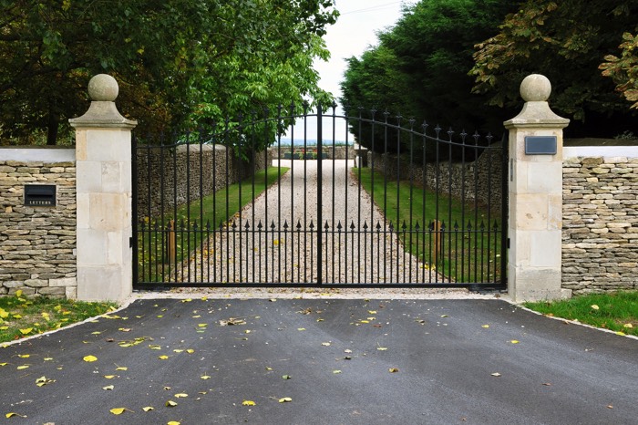 Entrée portail à double battants avec deux chapeaux en pierres. Mur en pierres avec allée gravillonnée, pelouse et grands arbres.