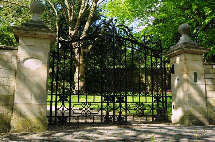 Entré portail à deux battants avec deux chapeaux en pierre. Portail de couleur noir avec allée en pavés, jardin et grands arbres.
