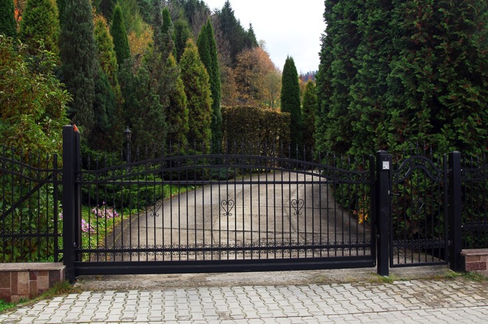 Entrée de portail avec barrettes verticales en fer forgé. Portail à double battants avec allée goudronnée et entourée de sapins et d'arbres différents.