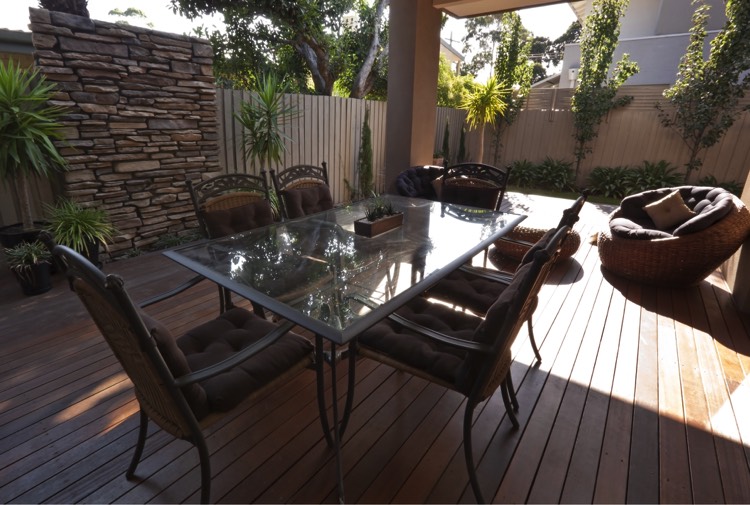 Aménagement extérieur de la maison avec tonnelle abritant une table à manger rectangulaire en verre sur parquet avec chaises. Pouf marron sur le côté. idée déco pour aménager l'extérieur de sa maison avec brio