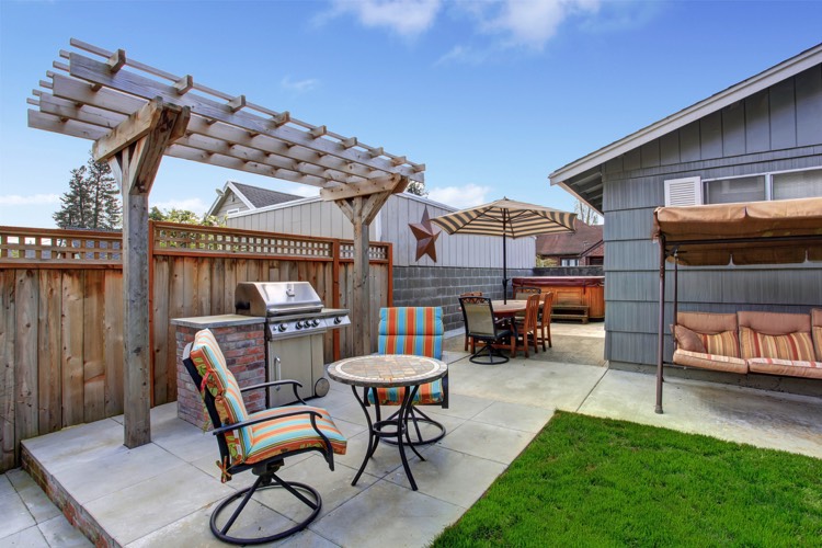 Aménagement d'un extérieur d'une maison avec petite tonnelle en bois. Carrelage, pelouse, parasol, petite table ronde et fauteuils style pop oranges et bleus.