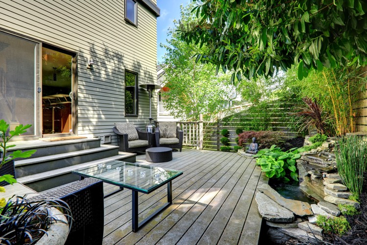 Aménagement d'un extérieur maison avec baie vitrée et parquet. Petite table en verre avec fauteuil et espace green power zen avec mini lac.