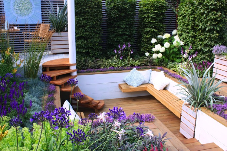 Aménagement d'un extérieur d'une maison avec banc en L en bois cerisier et parquet. Jardin avec mur végétal avec lierre, plantes blanches et violettes.