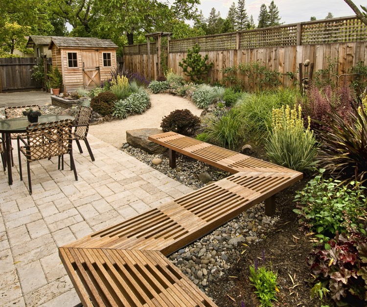 Aménager un extérieur avec clôture, banc et petite cabane en bois hêtre. Allée de terre amenant vers une table ronde avec sièges tressés dans une végétation abondante.