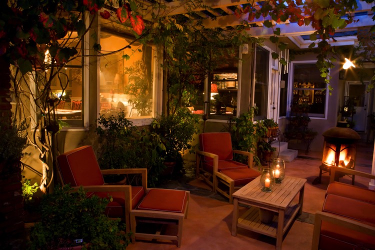 Extérieur maison green power avec pergola et plantes grimpantes. Petit salon avec table en bois, trois transats rouges et luminaires.