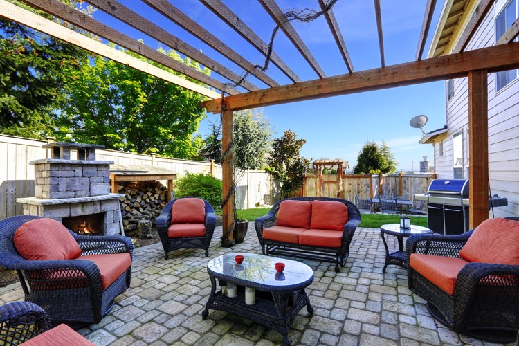 Aménagement extérieur de la maison avec pergola en bois chêne. Petit salon avec mini table en verre et fauteuils tressés oranges. Foyer extérieur en pierre avec stock de bois.