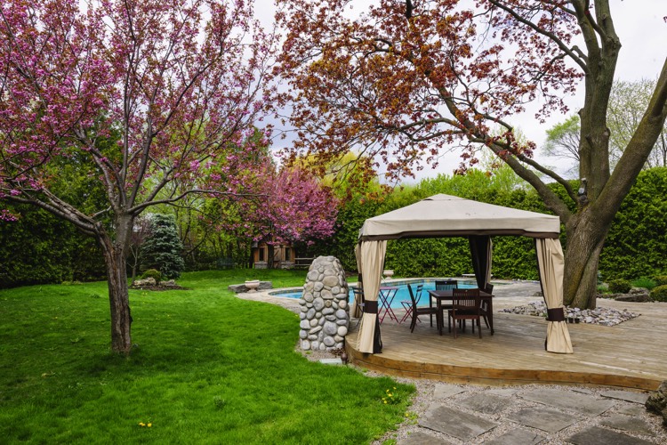 Aménagement d'un extérieur avec cerisier à fleurs et pelouse. Piscine avec margelles, terrasse avec parquet et salon abritée par une tonnelle.