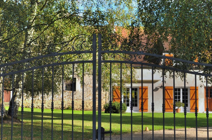 Portail en fer à rayures gris foncé en forme arrondie Grand jardin avec arbres et maison blanche et en pierres avec volets en bois cerisier.