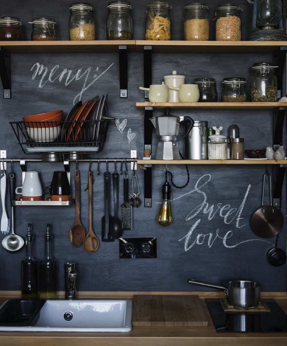 Cuisine tout en nuances de gris avec étagères en bois et multitude de différents pots en verre dessus. Ustensiles de cuisine accrochés et inscriptions blanches au mur.