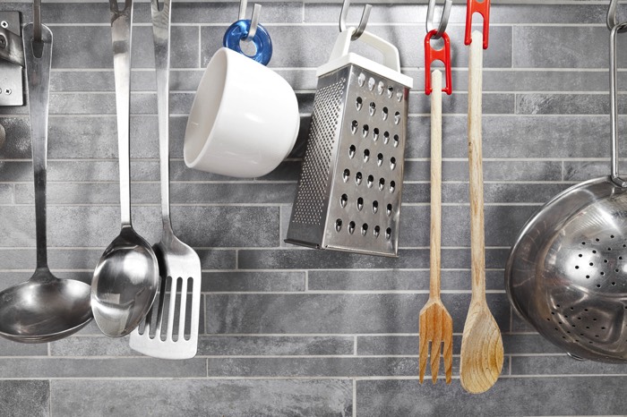Cuisine grise avec carrelage mural en gris clair. Multitude d'accessoires différents suspendus comme une louche, une cuillère, une tasse blanche ou encore une passoire. carrelage gris sur la crédence avec ustensiles de cuisine