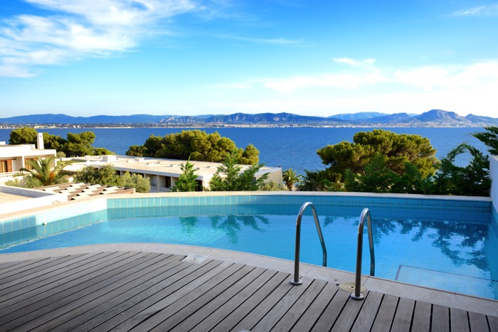 Aménager une piscine arrondie avec escalier et eau bleue, parquet tout autour et différents arbres à proximité. Magnifique panorama sur la mer.